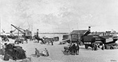 Stukgoederenoverslag bij Arnhem, 1929. Het vervoer van stukgoederen werd grotendeels verzorgd door de beurtvaart. Langs de kade zijn twee stoomboten afgemeerd om de goederen aan- en af te voeren. Bij het laden en lossen werd al van kleine hijskranen gebruikgemaakt. De aan- en afvoer over land geschiedt gedeeltelijk met vrachtauto’s, maar vaak ook nog met paard-en-wagens.