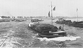 Druk binnenvaartverkeer in de scherpe Rijnbocht bij Stürzelberg in Duitsland, circa 1955. Op de voorgrond in de opvaart het kleine, geladen motorschip Cornelia dat aan de voormast de oploopvlag heeft gehesen, als teken dat het de voorgaande schepen wil passeren. Tegelijkertijd is aan de stuurhut de blauwe vlag uitgestoken, als teken dat het vaartuig de bakboordzijde (linkerzijde) van het vaarwater aanhoudt. Daardoor vaart het schip door de binnenbocht, waar de stroming van de rivier veel minder is dan in de buitenbocht. Aan de linkerzijde van de foto een afvarende sleep, voortgetrokken door een stoomsleepboot.